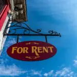Red and yellow 'For Rent' sign hanging from a building with a clear blue sky behind it.