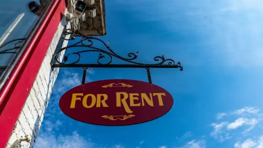 Red and yellow 'For Rent' sign hanging from a building with a clear blue sky behind it.
