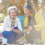Elderly mother and adult daughter sitting on a bench looking at their phones and laughing