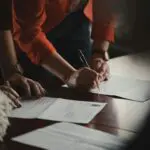 Close-up of two people signing paperwork at a desk.